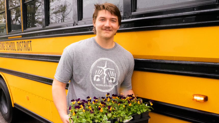 Bo Poljan C22 leans against a yellow school bus holding a flat of flowers during Work-A-Thon 2022
